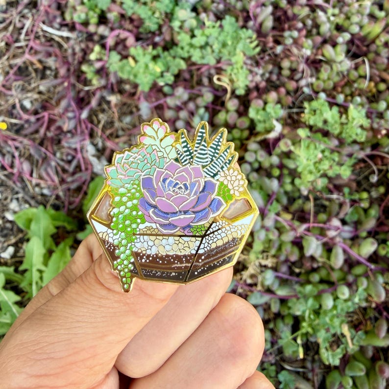May include: A gold-coloured enamel pin depicting a terrarium with succulents. The pin features a central purple succulent surrounded by green and white plants. The terrarium has a layered brown and white base.