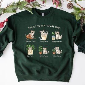 May include: Dark green sweatshirt with the text "THING I DO IN MY SPARE TIME" and illustrations of cats with plants. The illustrations show cats rearranging, repotting, and propagating plants, buying plants, talking to plants, and watering plants.