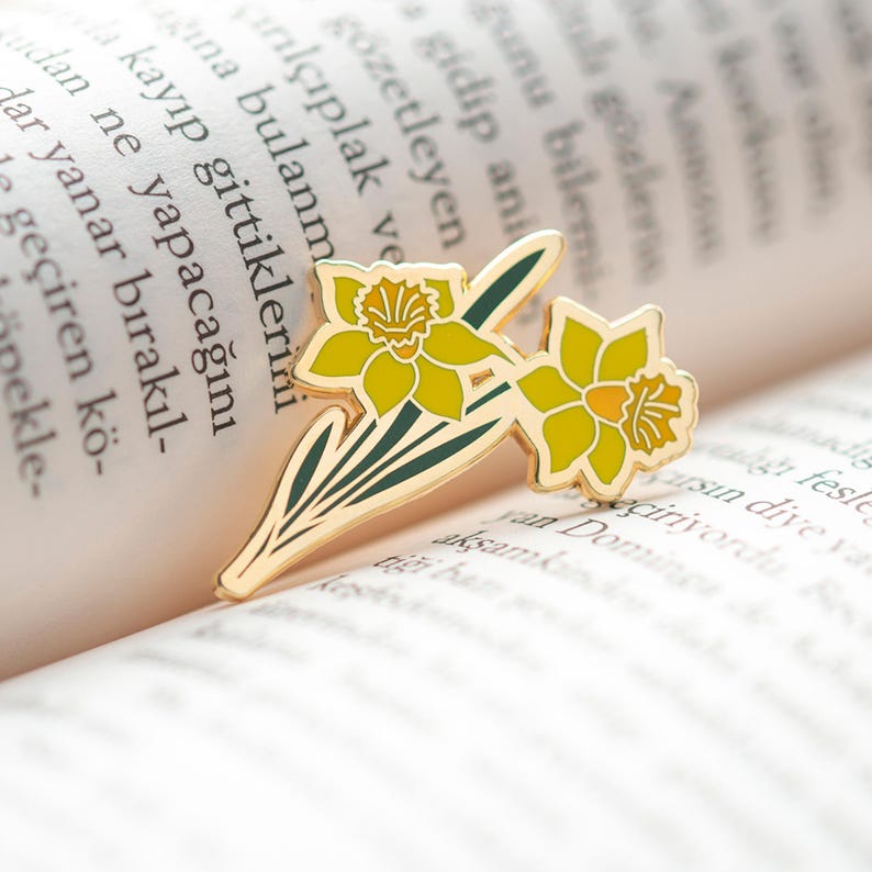 May include: Gold-tone enamel pin featuring two yellow daffodils with green stems and leaves. The pin is on a book.