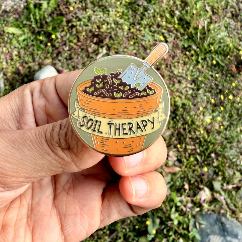 May include: A round enamel pin featuring a gardening theme. The pin depicts a terracotta pot filled with soil, sprouts, and a small trowel. The words "SOIL THERAPY" are written on a banner across the pot. The pin is held in a hand against a background of green grass.