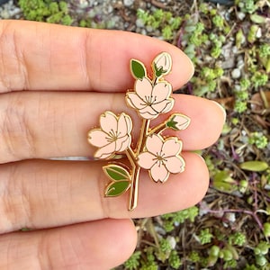 Op de afbeelding: Een goudkleurige metalen pin met een takje kersenbloesem. De pin heeft drie open roze bloemen, twee knoppen en groene bladeren. De bloesems hebben witte details en zijn gezet tegen een gouden omtrek.
