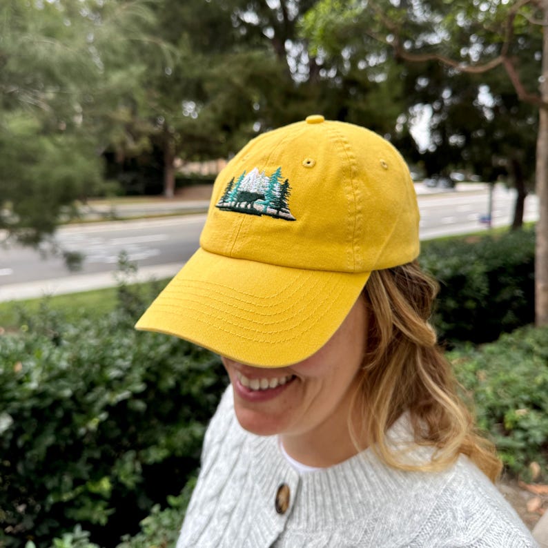 Puede incluir: Una gorra de b&eacute;isbol amarilla con un dise&ntilde;o bordado de una cordillera y &aacute;rboles. La gorra tiene una visera curvada y una correa ajustable.