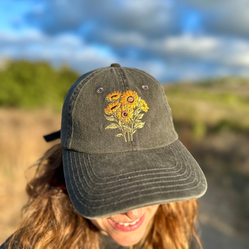 May include: A dark gray baseball cap with an embroidered design of yellow sunflowers. The cap has a curved brim and a structured crown.