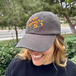 Puede incluir: Una gorra de béisbol gris con un diseño bordado de amapolas de California naranjas.
