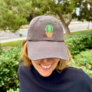 Puede incluir: Una gorra de béisbol gris con el texto "Pot Head" bordado en la parte delantera. Un cactus verde con flores rojas está bordado junto al texto.