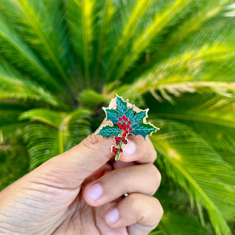 May include: Gold-tone enamel pin featuring a sprig of holly with green leaves and red berries.
