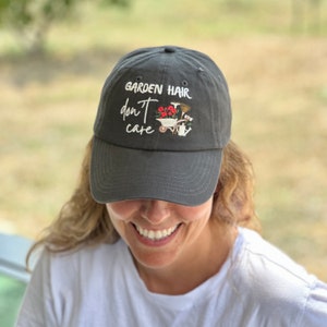 Op de afbeelding: Een donkergrijze baseballcap met de geborduurde tekst "GARDEN HAIR don't care" in witte letters. De cap heeft een klein geborduurd ontwerp van een kruiwagen met bloemen en tuingereedschap. De hoed wordt door een persoon gedragen.