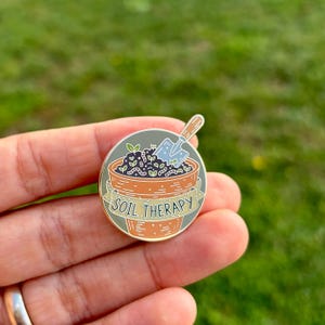 May include: A round enamel pin featuring a terracotta-colored flower pot with the words "SOIL THERAPY" on a banner. The pot contains soil, plants, and a small blue shovel. The pin is held in a hand against a blurred green background.