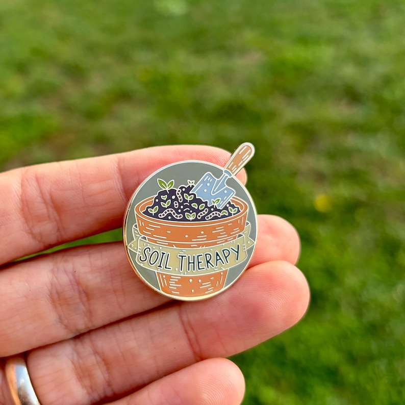 May include: A round enamel pin featuring a terracotta-colored flower pot with the words "SOIL THERAPY" on a banner. The pot contains soil, plants, and a small blue shovel. The pin is held in a hand against a blurred green background.
