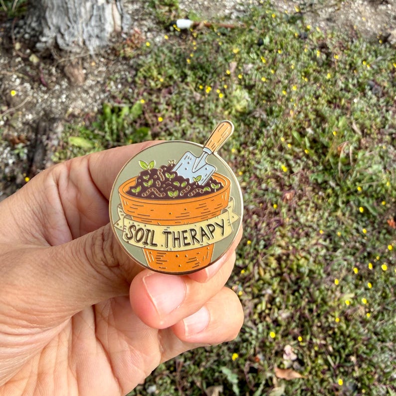 May include: A round enamel pin featuring a terracotta-colored flower pot filled with soil, sprouts, and a small trowel. The pin has a banner that reads "SOIL THERAPY". The pin is held in a hand against a background of green foliage.