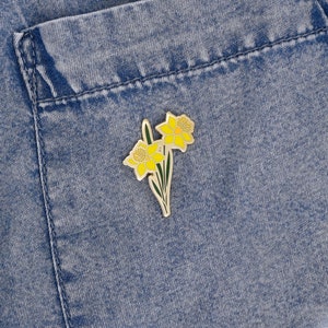 May include: A gold-colored enamel pin featuring two yellow daffodils with green stems and leaves. The pin is attached to a denim fabric background.