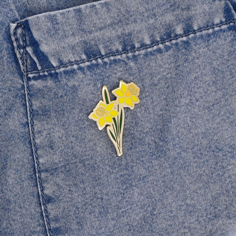 May include: A gold-colored enamel pin featuring two yellow daffodils with green stems and leaves. The pin is attached to a denim fabric background.