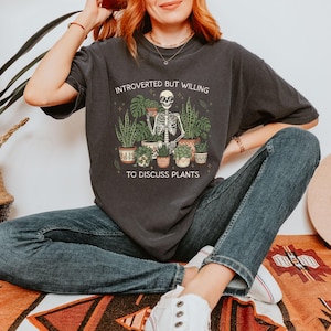 May include: Dark gray t-shirt with the text "INTROVERTED BUT WILLING TO DISCUSS PLANTS" and a graphic of a skeleton surrounded by potted plants. The person is wearing blue jeans and white sneakers.