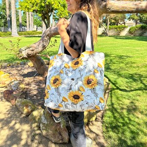 K&ouml;nnte beinhalten: Eine wei&szlig;e Einkaufstasche mit einem Sonnenblumenmuster. Die Tasche hat eine wei&szlig;e Canvas-Basis mit einem floralen Muster aus Sonnenblumen und Bl&auml;ttern in Gelb- und Gr&uuml;nt&ouml;nen.