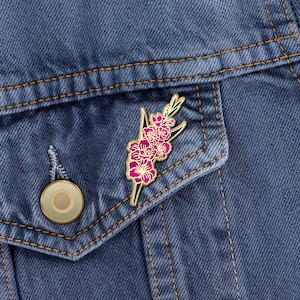 May include: A gold-toned enamel pin featuring a pink flower with green leaves. The pin is attached to a denim jacket.
