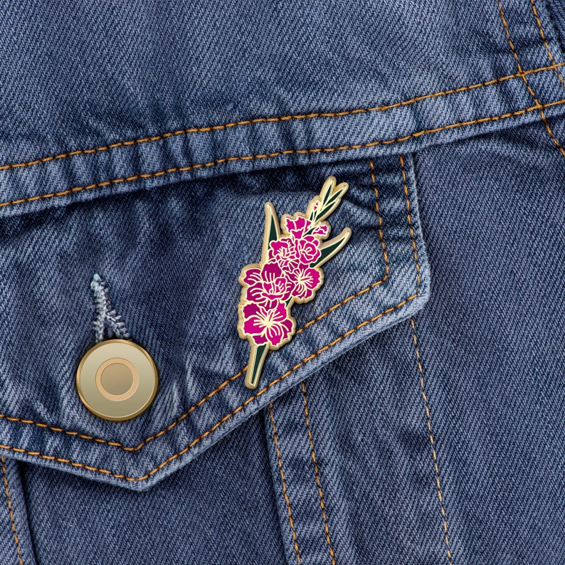 May include: A gold-toned enamel pin featuring a pink flower with green leaves. The pin is attached to a denim jacket.