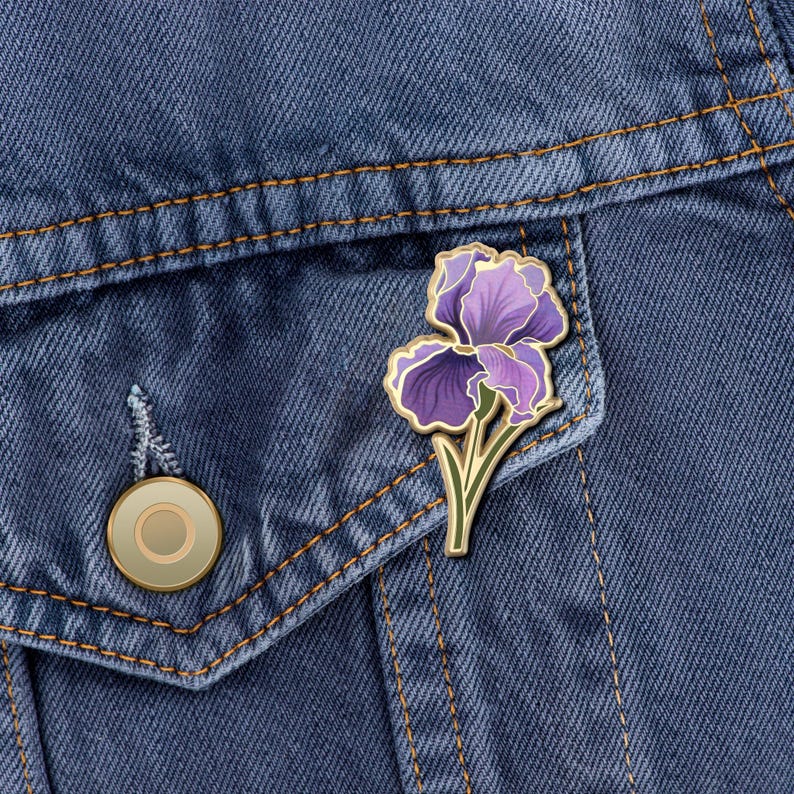 May include: A purple and gold enamel pin featuring a stylized iris flower. The pin is attached to a denim jacket.