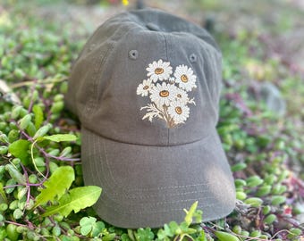 May include: A dark gray baseball cap with an embroidered floral design. The embroidery features a cluster of white daisies with yellow centers and brown stems. The cap is made of a soft fabric and has a curved brim.