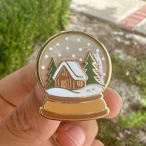 May include: A gold-colored enamel pin in the shape of a snow globe. Inside the globe is a small house with a white roof, two green trees, and white snow. The base of the snow globe is brown and gold.