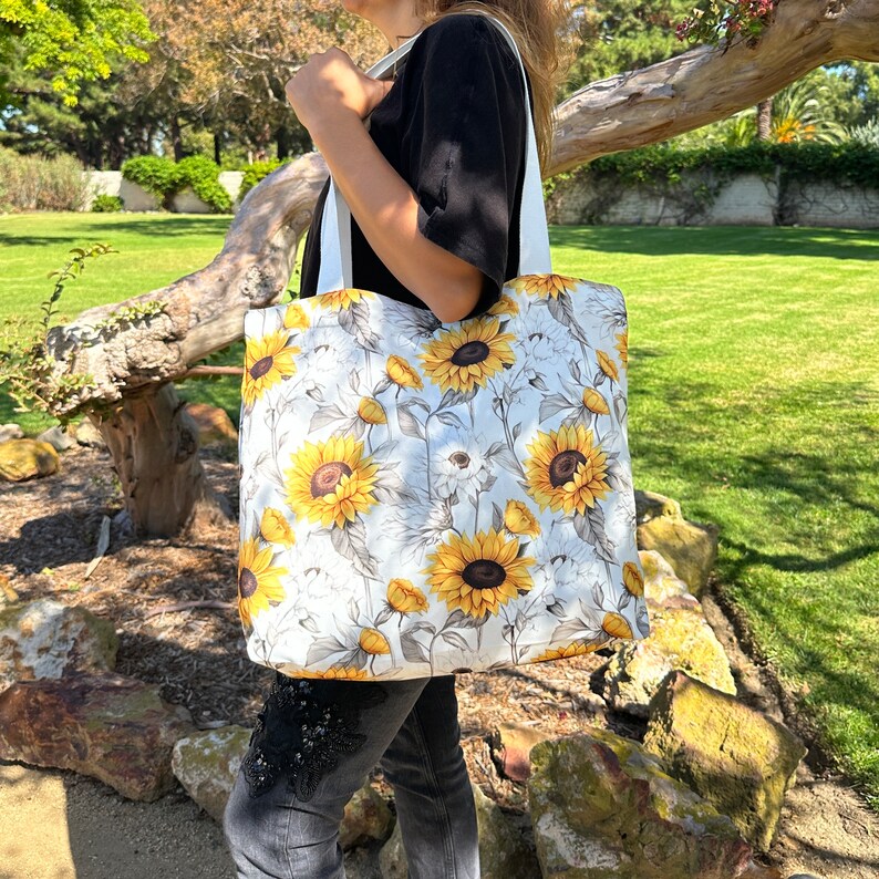 K&ouml;nnte beinhalten: Eine wei&szlig;e Einkaufstasche mit einem Sonnenblumenmuster. Die Tasche hat wei&szlig;e Henkel und ist aus Canvas.