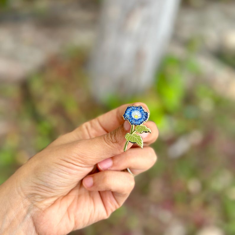 May include: Gold-tone enamel pin featuring a blue morning glory flower with a white center and green leaves.
