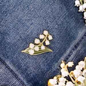 May include: A gold-tone enamel pin featuring a white lily of the valley flower with green leaves. The pin is on a blue denim background.