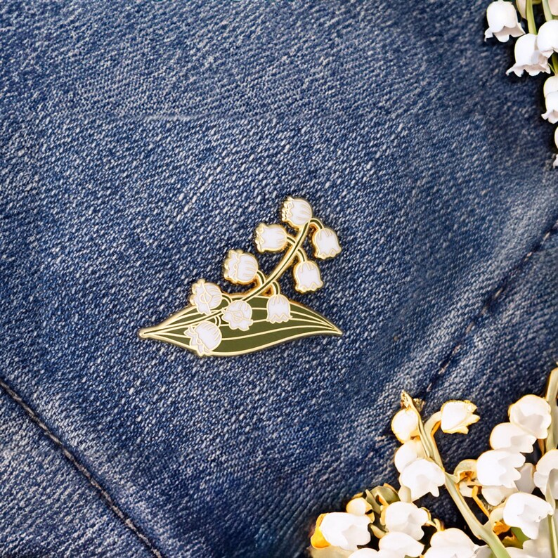 May include: A gold-tone enamel pin featuring a white lily of the valley flower with green leaves. The pin is on a blue denim background.