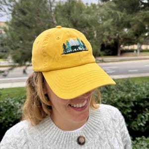 Puede incluir: Una gorra de b&eacute;isbol amarilla con un dise&ntilde;o bordado de una cordillera con &aacute;rboles y un oso. El dise&ntilde;o es en verde, blanco y negro.