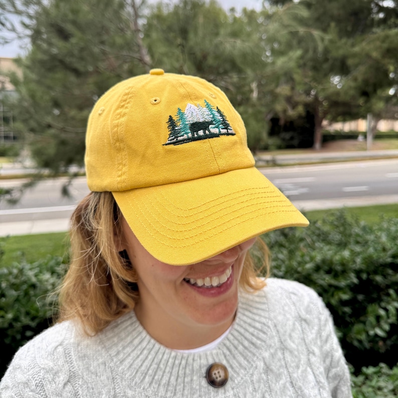 Puede incluir: Una gorra de b&eacute;isbol amarilla con un dise&ntilde;o bordado de una cordillera con &aacute;rboles y un oso. El dise&ntilde;o es en verde, blanco y negro.