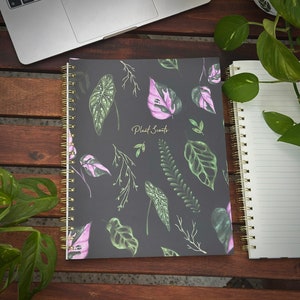 Könnte beinhalten: Ein schwarzes Spiralnotizbuch mit einem botanischen Druck mit rosa und grünen Blättern. Der Notizbuchumschlag trägt den Text "Plant Scouts" in goldenen Buchstaben.