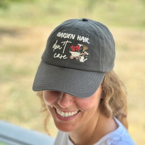 Op de afbeelding: Een grijze baseballcap met de geborduurde tekst "GARDEN HAIR don't care". De cap heeft een klein geborduurd ontwerp van tuingereedschap en bloemen. De hoed is gemaakt van een zacht materiaal en heeft een gebogen klep.