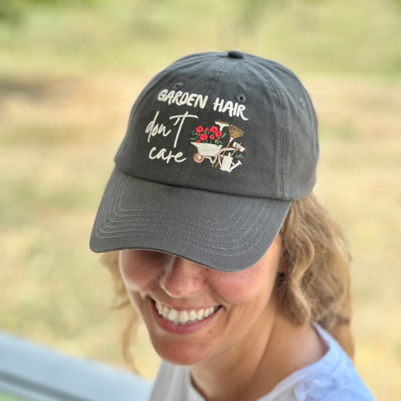 Op de afbeelding: Een grijze baseballcap met de geborduurde tekst "GARDEN HAIR don't care". De cap heeft een klein geborduurd ontwerp van tuingereedschap en bloemen. De hoed is gemaakt van een zacht materiaal en heeft een gebogen klep.
