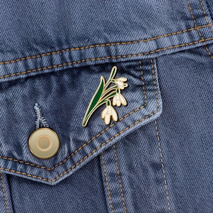 May include: A gold enamel pin featuring a white flower with green leaves. The pin is on a blue denim jacket.