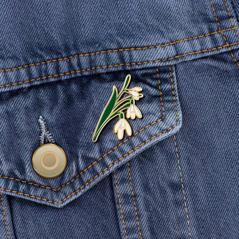 May include: A gold enamel pin featuring a white flower with green leaves. The pin is on a blue denim jacket.