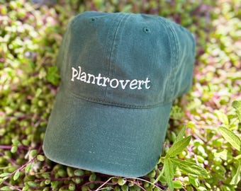 May include: A dark green baseball cap with the word "plantrovert" embroidered in white. The hat is resting on a bed of green and purple plants. The cap has a curved brim and a classic six-panel design.