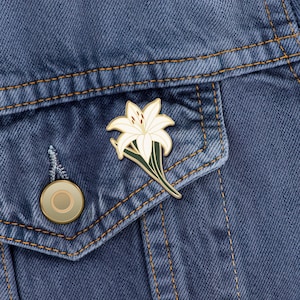 May include: A white enamel pin of a stylized white lily flower with green leaves on a denim jacket.