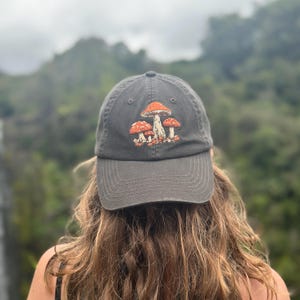 Könnte beinhalten: Dunkelgraue Baseballkappe mit einem gestickten Design aus drei orangefarbenen und weißen Pilzen. Die Kappe hat einen gebogenen Schirm und ein klassisches Sechs-Panel-Design. Der Hintergrund ist verschwommen, mit einer natürlichen Umgebung im Freien.