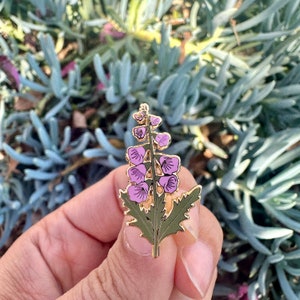 Può includere: Spilla in smalto dorata con un fiore viola con foglie verdi. Il fiore ha una forma a campana ed è composto da più petali.