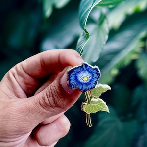 May include: A gold-tone enamel pin featuring a blue flower with a yellow center and green leaves. The pin is held in a hand against a green background.