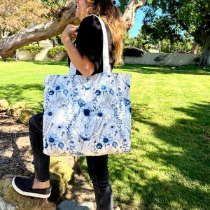 Op de afbeelding: Een witte tote bag met een blauw bloemenpatroon. De tas heeft witte banden en wordt door een persoon vastgehouden.