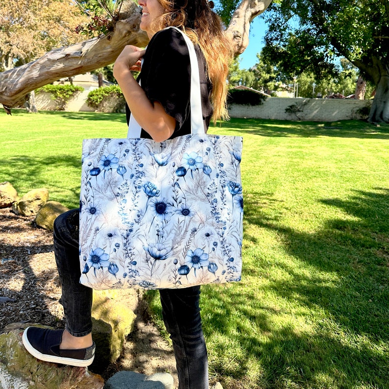 Op de afbeelding: Een witte tote bag met een blauw bloemenpatroon. De tas heeft witte banden en wordt door een persoon vastgehouden.