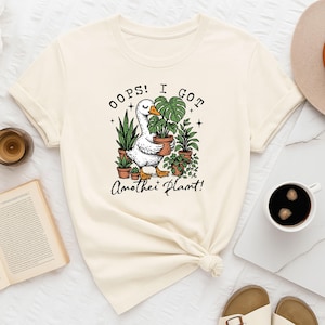Könnte beinhalten: Cremefarbenes T-Shirt mit einer humorvollen Illustration einer weißen Gans, die eine Topfpflanze hält, umgeben von anderen Topfpflanzen. Der Text "Oops! I got Another Plant!" ist über und unter der Gans. Das T-Shirt ist an der Taille geknotet.