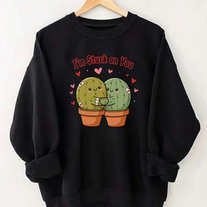 Może przedstawiać: Czarna bluza z napisem "I'm Stuck on You" nad kreskówkową ilustracją dwóch obejmujących się kaktusów w doniczkach. Kaktusy są zielone i mają bandaże. Rozrzucone są czerwone serca.