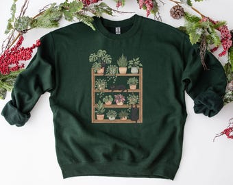 Può includere: Felpa verde scuro con una grafica di una mensola in legno piena di piante in vaso. Due gatti neri sono raffigurati sulla mensola. La felpa ha un girocollo e maniche lunghe. Il design utilizza tonalità di verde, marrone e rosa.