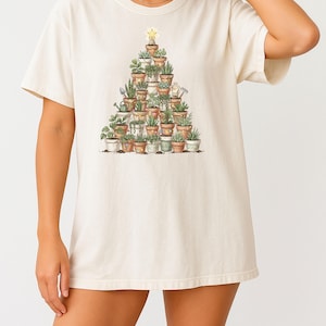 May include: Cream-colored t-shirt featuring a watercolor illustration of a Christmas tree made of potted plants. The tree is topped with a gold star. The shirt is a relaxed fit.