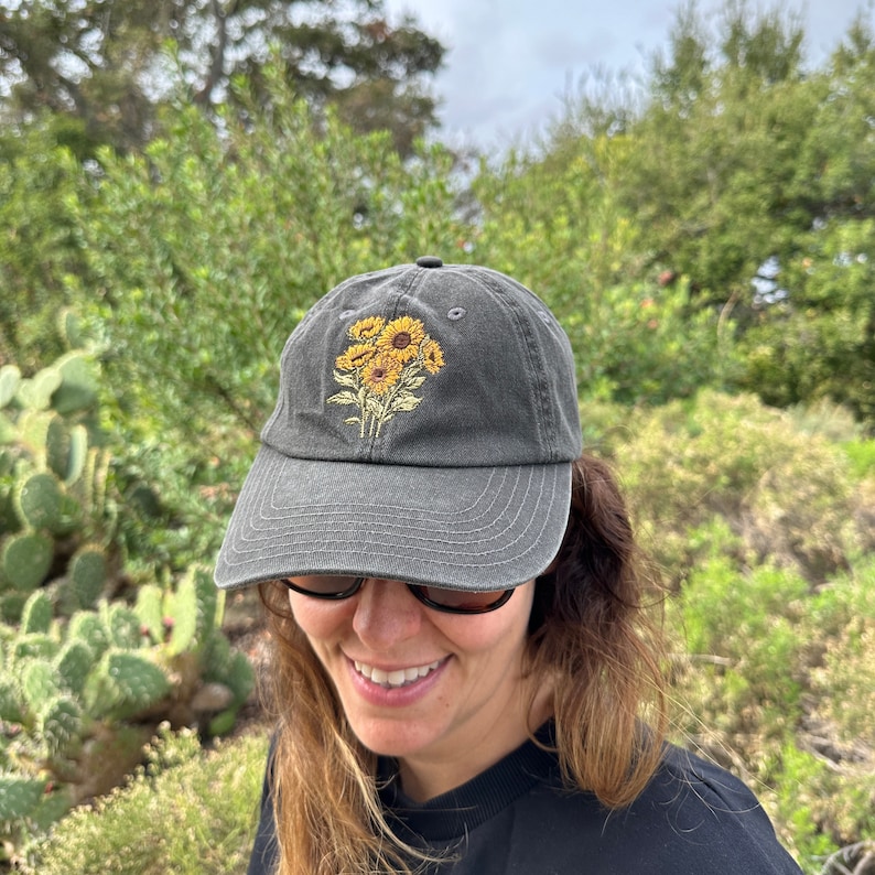 May include: A dark gray baseball cap with an embroidered design of yellow sunflowers. The cap has a curved brim and a structured crown.