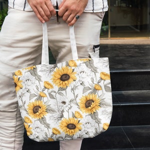 K&ouml;nnte beinhalten: Eine wei&szlig;e Canvas-Tragetasche mit einem Sonnenblumenmuster. Die Tasche hat zwei Henkel und wird von einer Person gehalten.