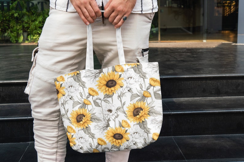 K&ouml;nnte beinhalten: Eine wei&szlig;e Canvas-Tragetasche mit einem Sonnenblumenmuster. Die Tasche hat zwei Henkel und wird von einer Person gehalten.