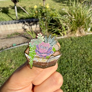Op de afbeelding: Een kleurrijke emaille pin met een terrariumontwerp met verschillende vetplanten. De pin toont een centrale paarse en roze bloem omringd door groene en witte planten, allemaal geplaatst in een geometrische, bruin getinte container.