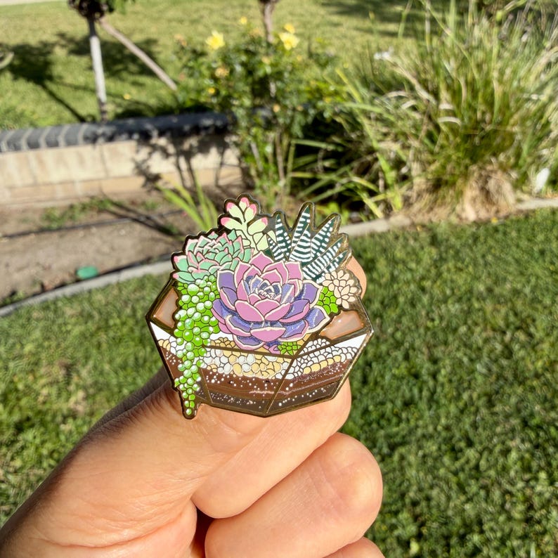 May include: A colourful enamel pin featuring a terrarium design with various succulents. The pin showcases a central purple and pink flower surrounded by green and white plants, all set within a geometric, brown-toned container.